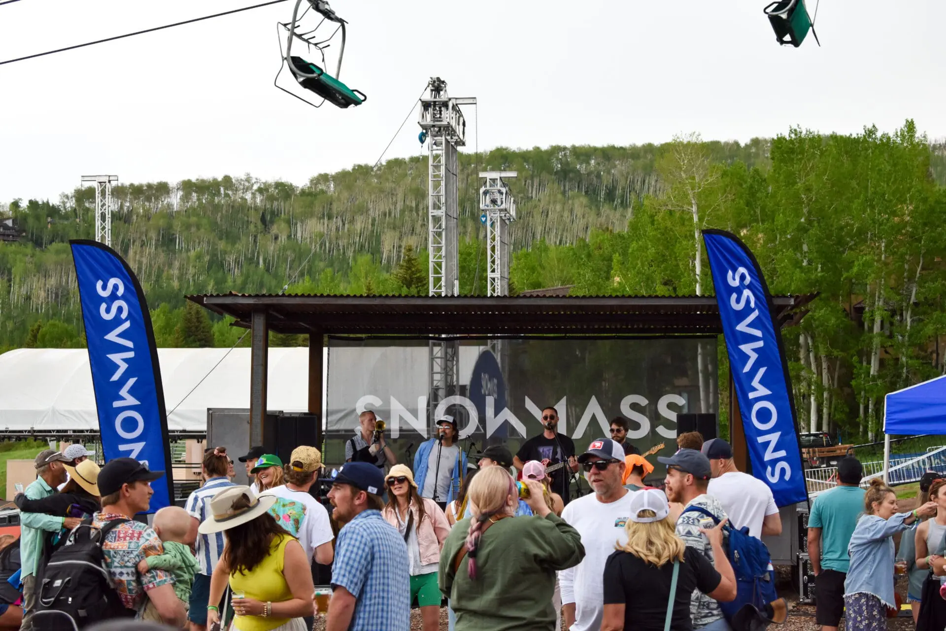 Rock Garden Concert Series on Fanny Hill at the end of the Snowmass Mall with the chairlift hanging above, and blue Snowmass flags among a big crowd of people.