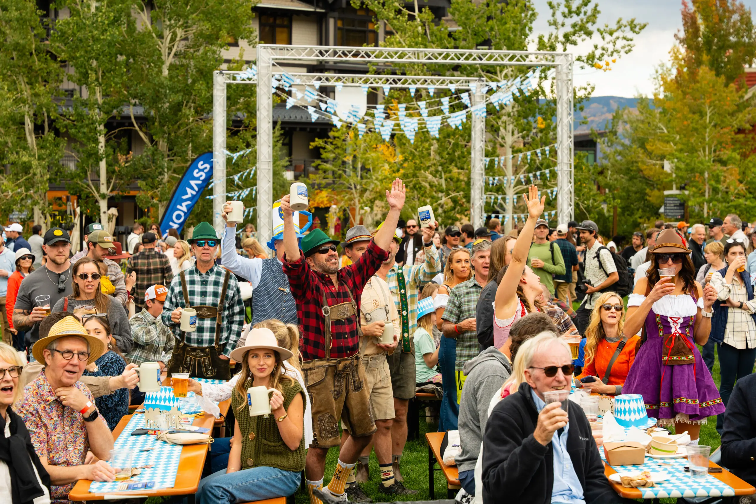 A crowd celebrates Snowmass Oktoberfest, with a man dressed in a red plaid shirt and lederhosen "prosts" on the lawn.