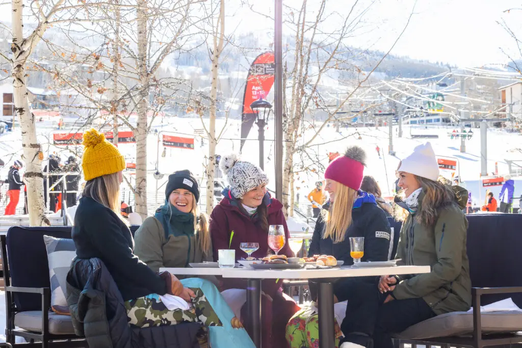 A group of four women enjoy spring in Snowmass with après in the sunshine, with Snowmass Ski Area in the background.