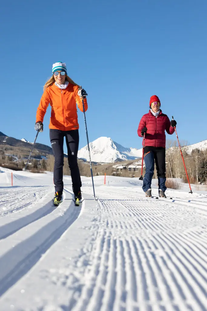 A woman in orange and a woman in red Nordic ski on the Snowmass course on a blue sky day, enjoying a Snowmass, Colorado winter vacation.