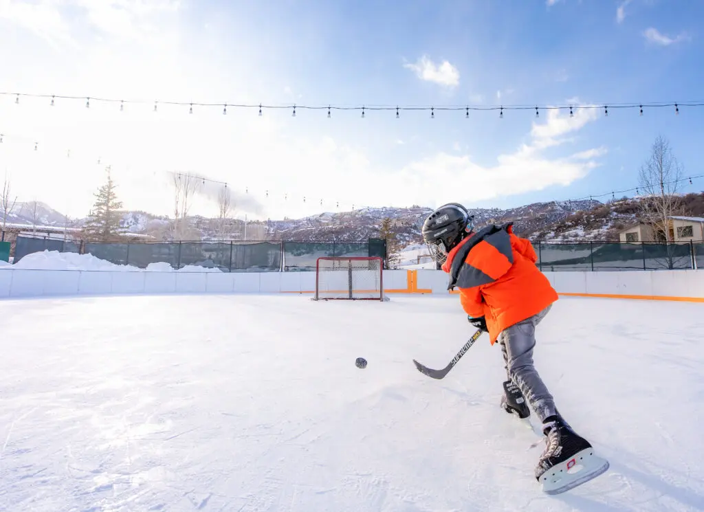 Boy in an orange jacket shoots a hockey puck into the net at the Snowmass Town Park ice skating rink,