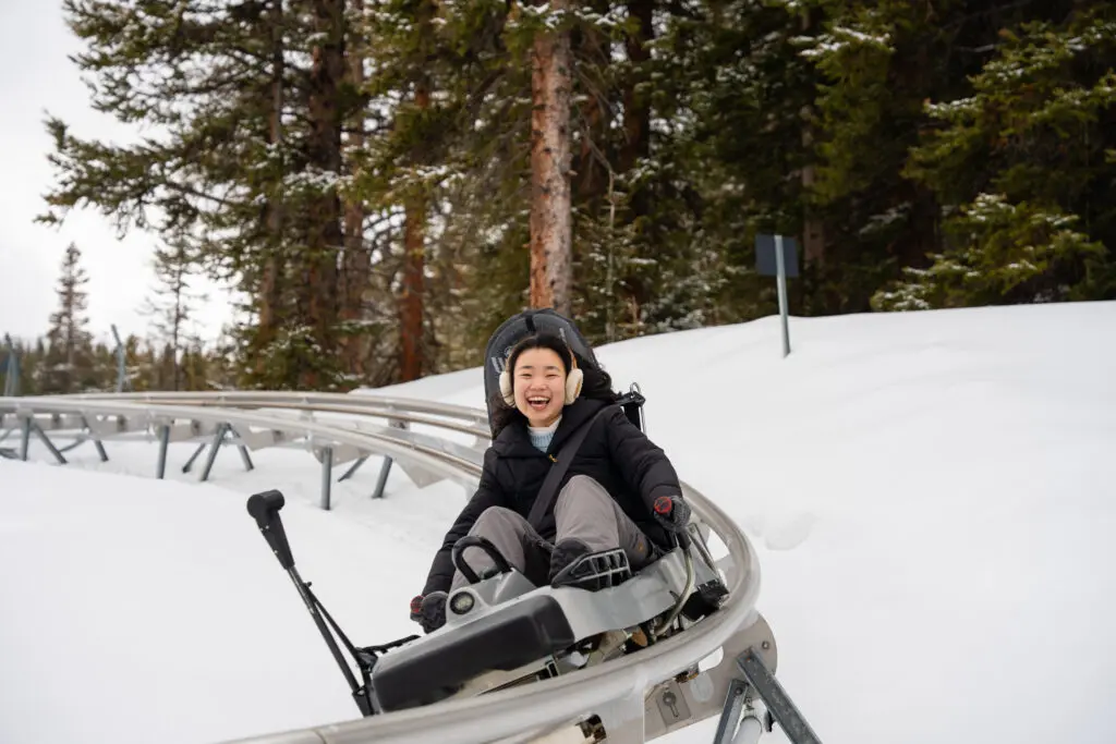 A girl rides the alpine coaster in Snowmass with a big smile on her face, wearing earmuffs while on a Snowmass, Colorado winter vacation.