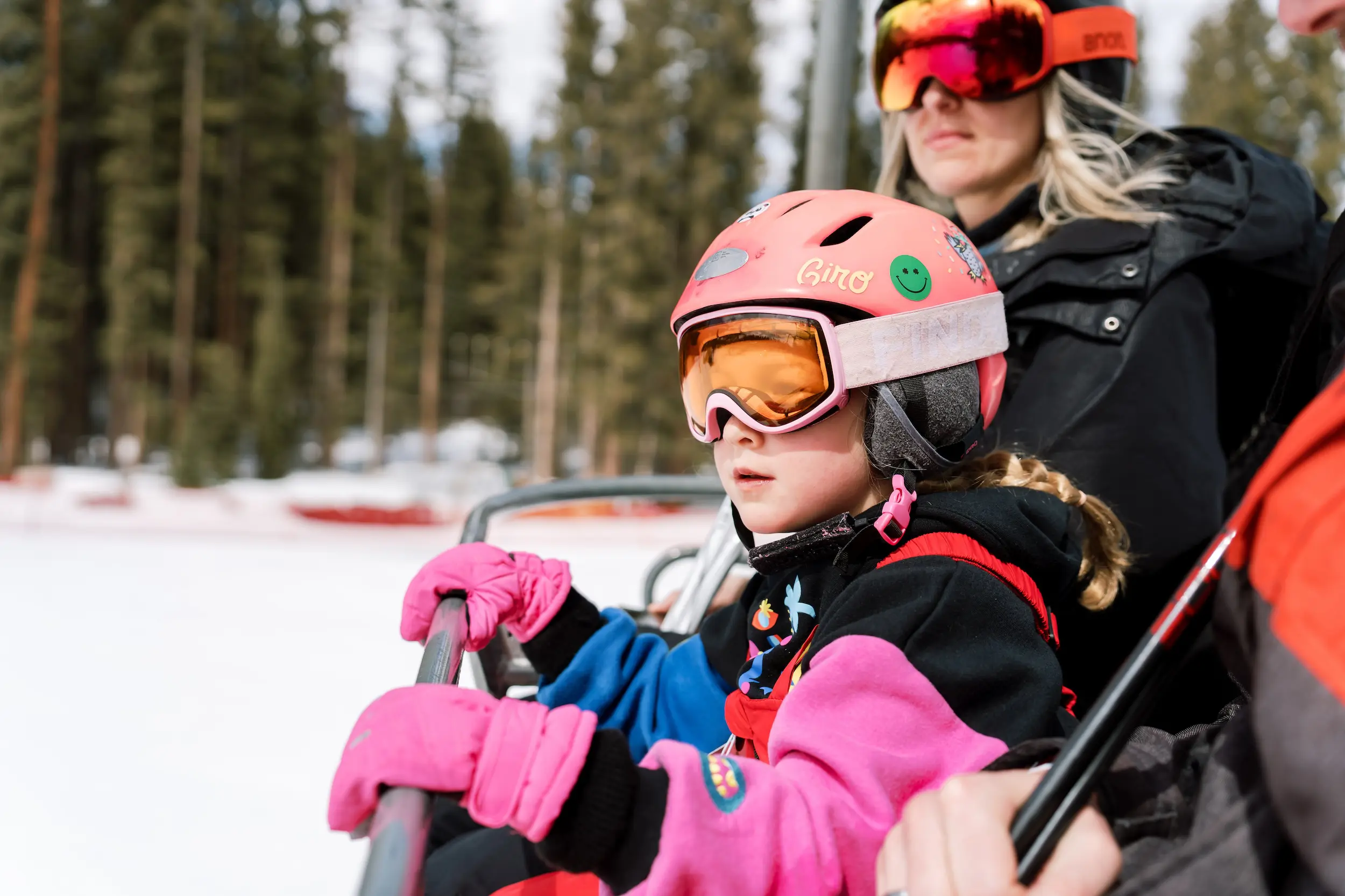 little girl wearing ski goggles and a helmet while on a chairlift with her mother