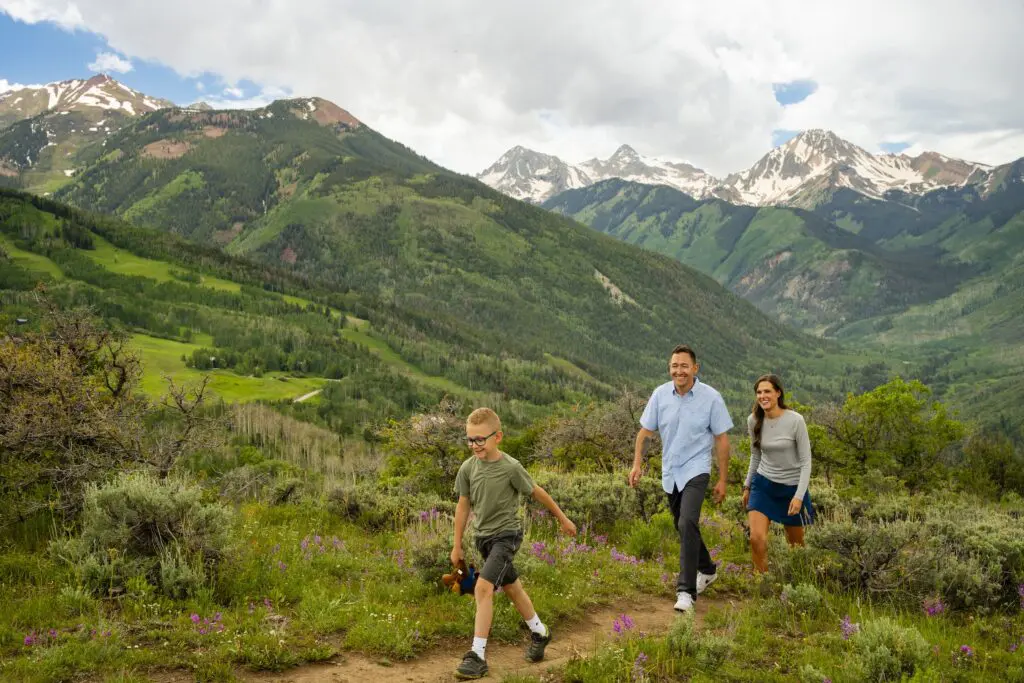 two adults and their child hiking on a summer trail in the mountains near Aspen Colorado with lightly snowy peaks in the background